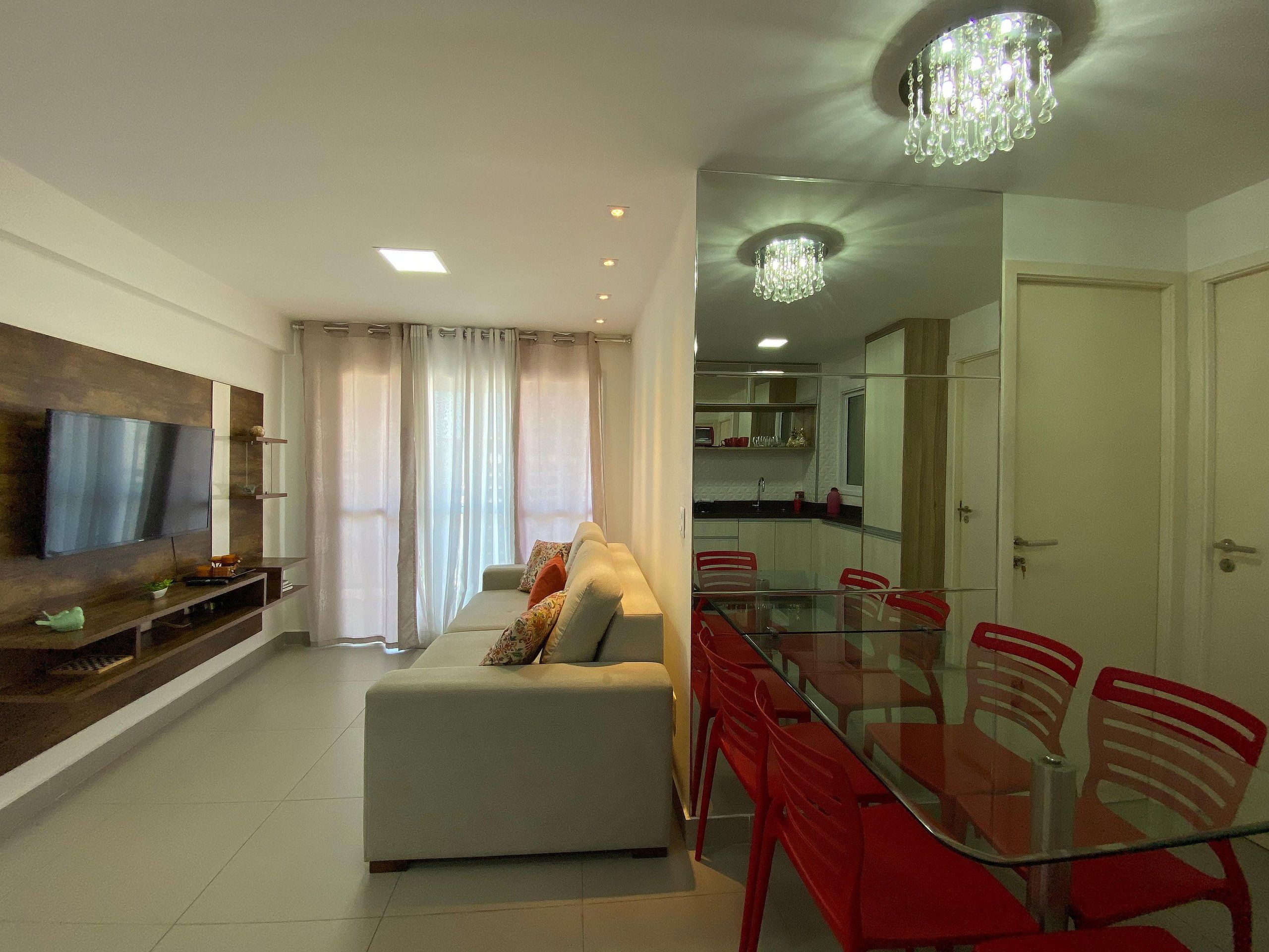 Una sala de estar moderna con televisor de pantalla plana, un sofá, una mesa de comedor de vidrio con sillas rojas y una zona de cocina.
