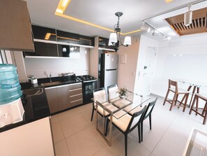 A modern kitchen with a gas stove, refrigerator, and a dining area with a glass table and wooden chairs. - Compacto de Luxo - Helbor My Way (Fortaleza)