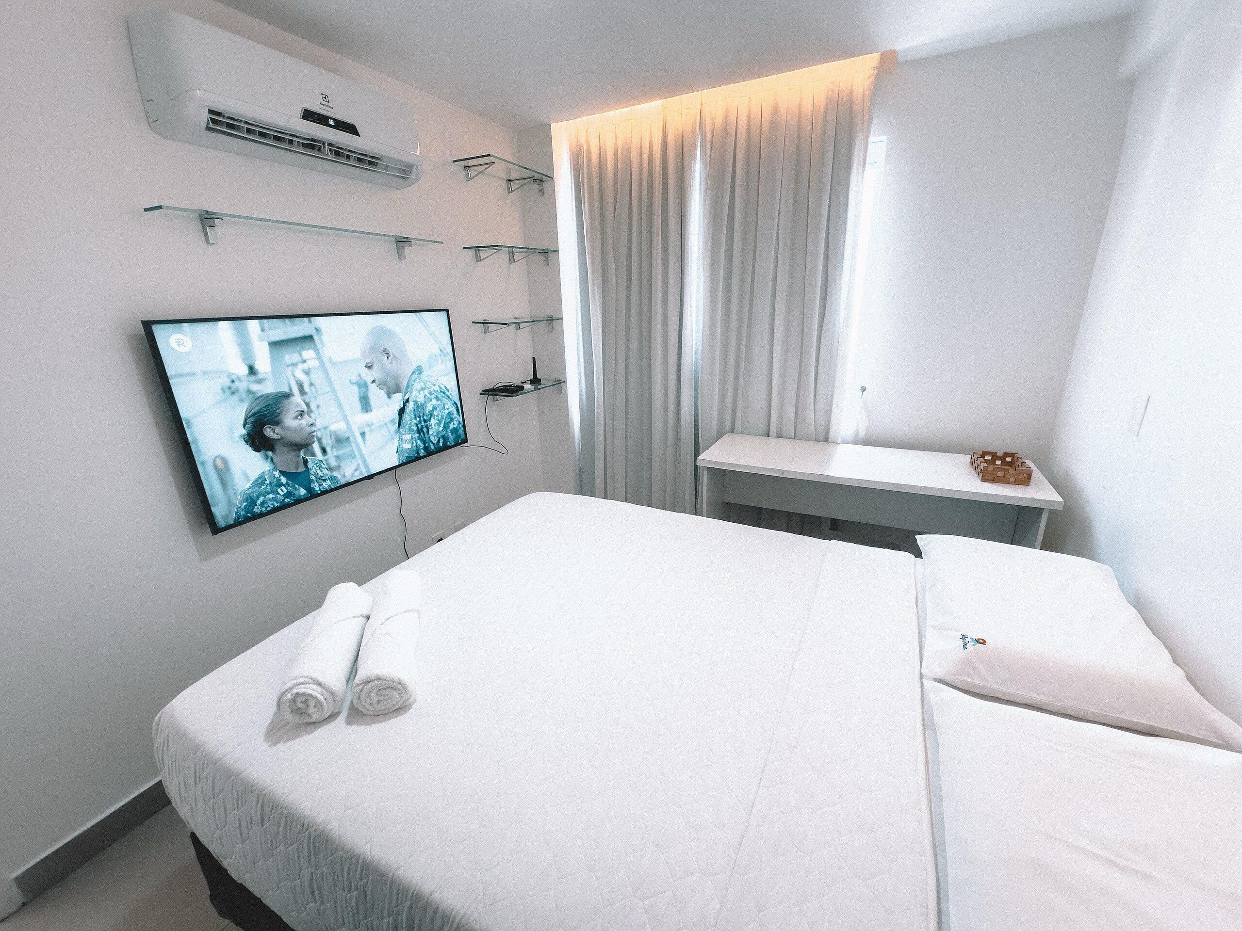Ett hotellrum med en säng, en tv som är monterad på väggen, ett litet bord och ett fönster med gardiner.