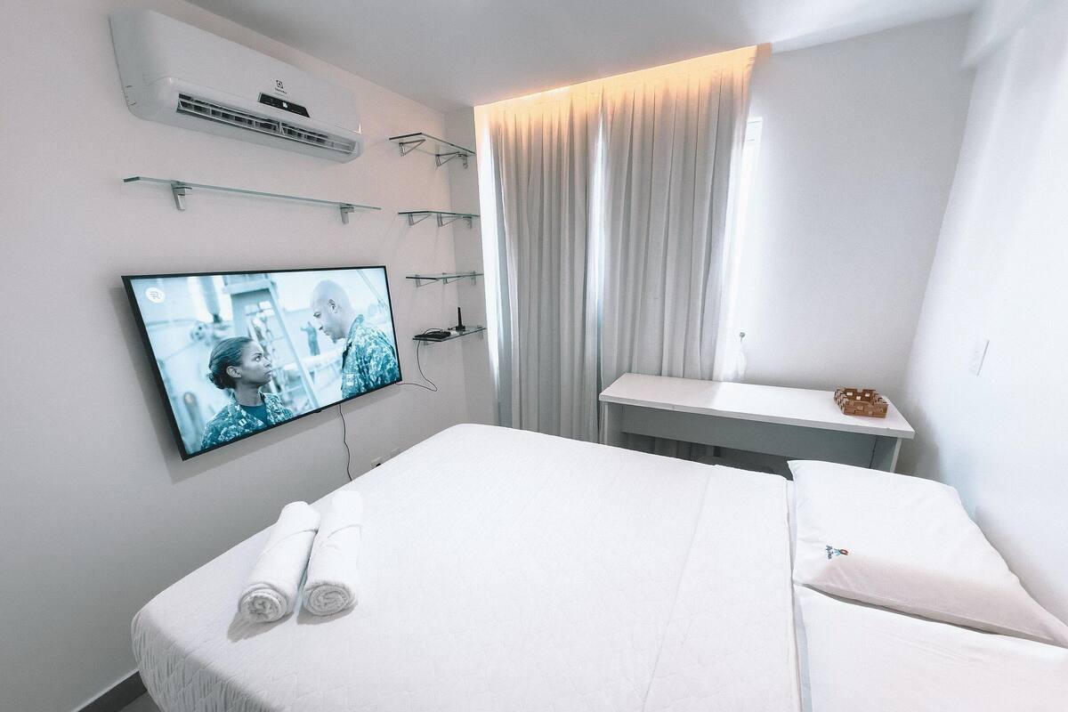 Ett hotellrum med en säng, en tv som är monterad på väggen, ett litet bord och ett fönster med gardiner.