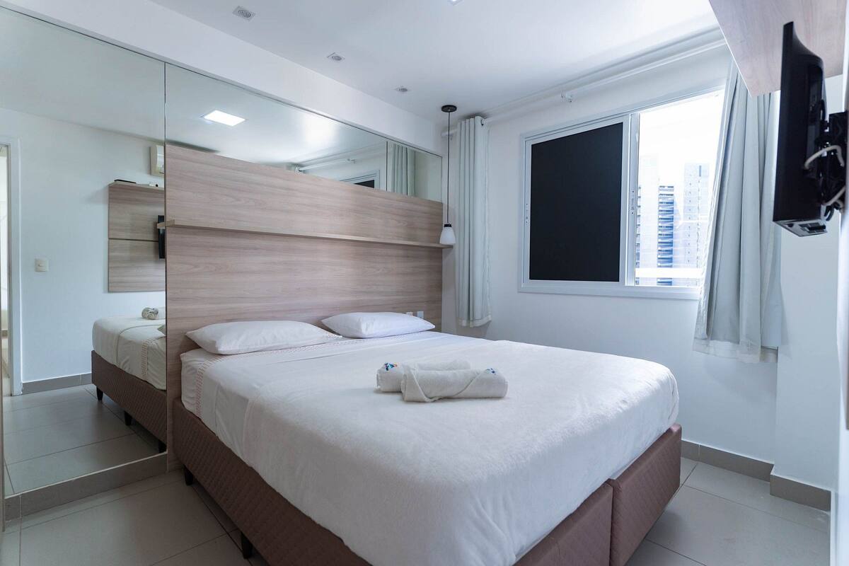 A modern hotel room with a large bed, a mirror, a TV, and a window with a city view.