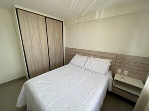 A neatly made bed with a wooden headboard, a bedside table with a lamp, and a wardrobe with a sliding door. - Compacto de Luxo - Helbor My Way (Fortaleza)