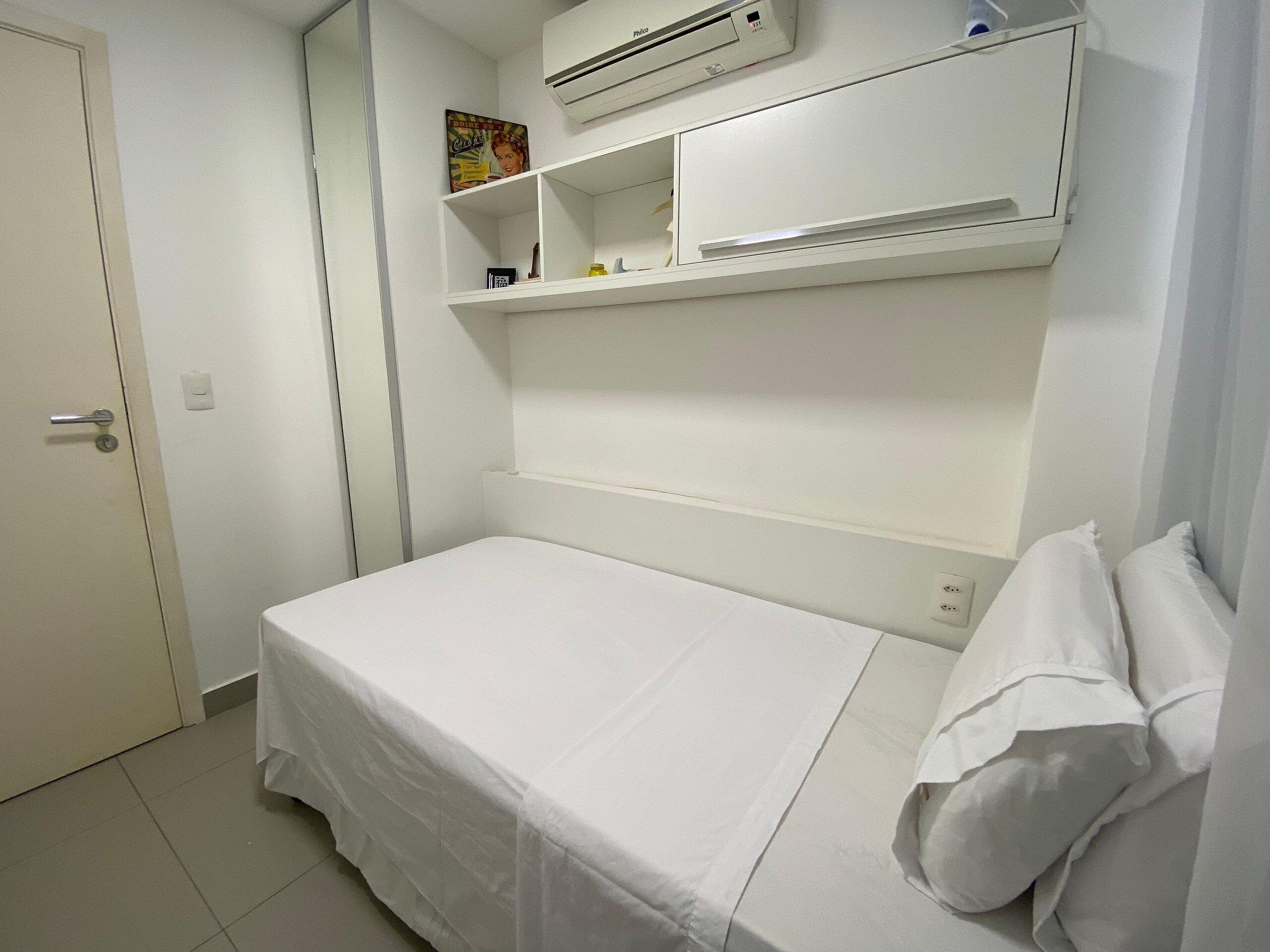 A small, white room with a single bed, a shelf with a few items, and an air conditioning unit above the bed.