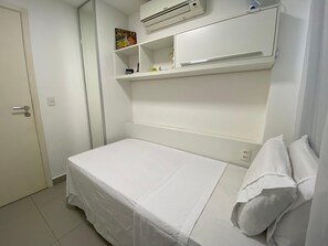 A small, white room with a single bed, a shelf with a few items, and an air conditioning unit above the bed. - Compacto de Luxo - Helbor My Way (Fortaleza)
