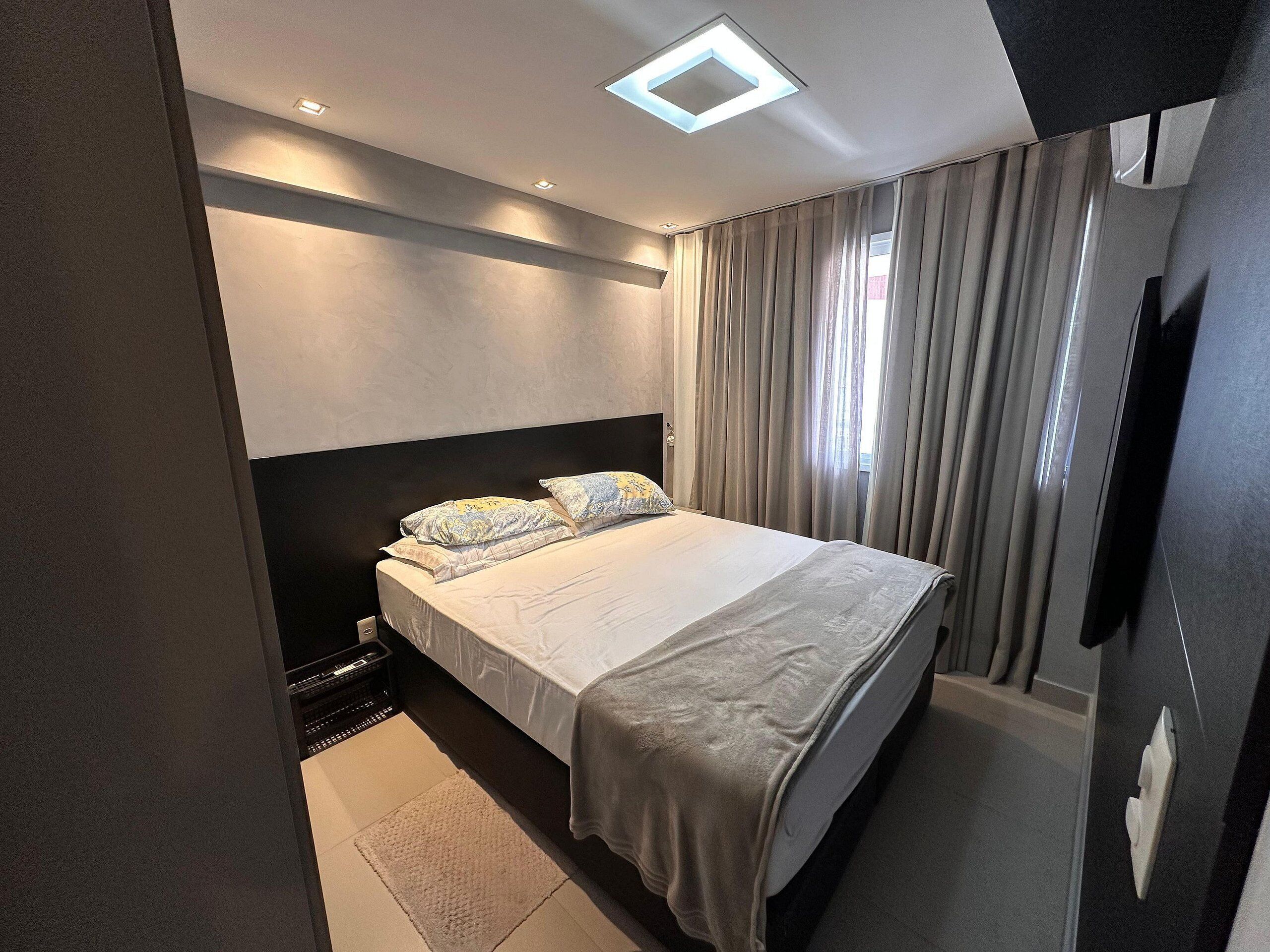 Un dormitorio moderno con cama, cortinas, una lámpara de techo y un televisor montado en la pared.