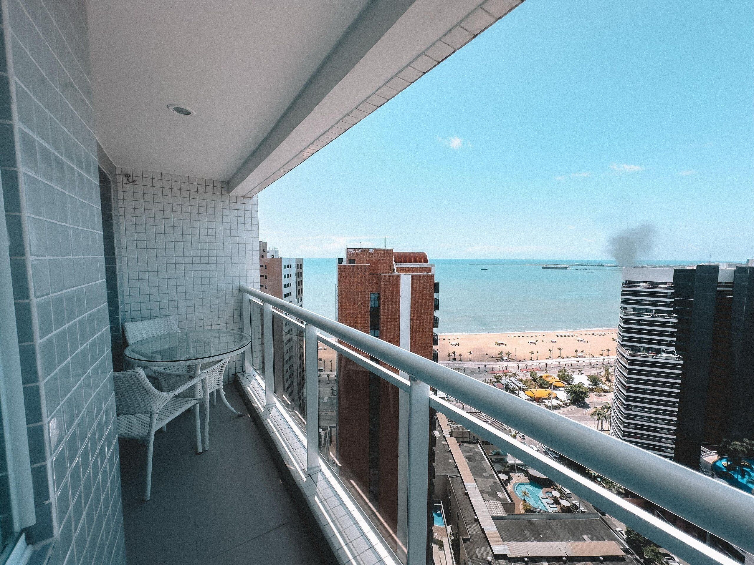 Un balcon avec une table et des chaises en verre, donnant sur une plage et une piscine.