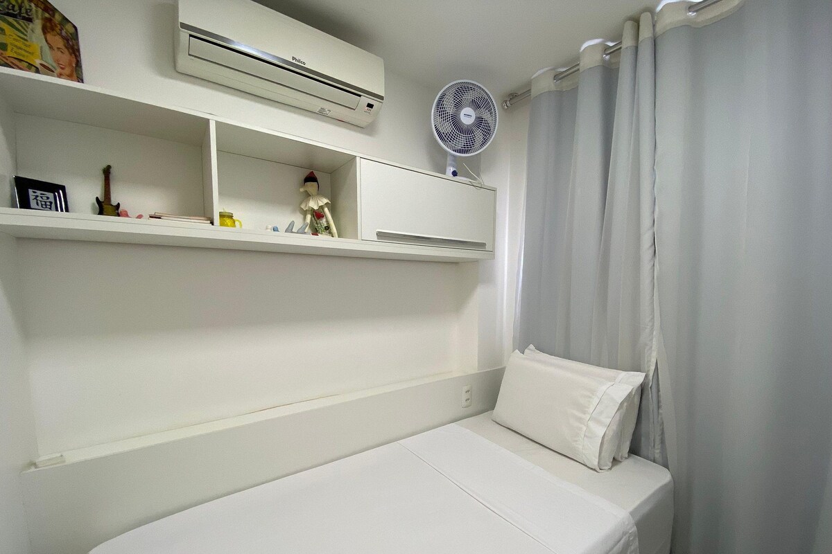 A small, neatly arranged bedroom with a single bed, a shelf with personal items, a wall-mounted air conditioner, and a curtain.