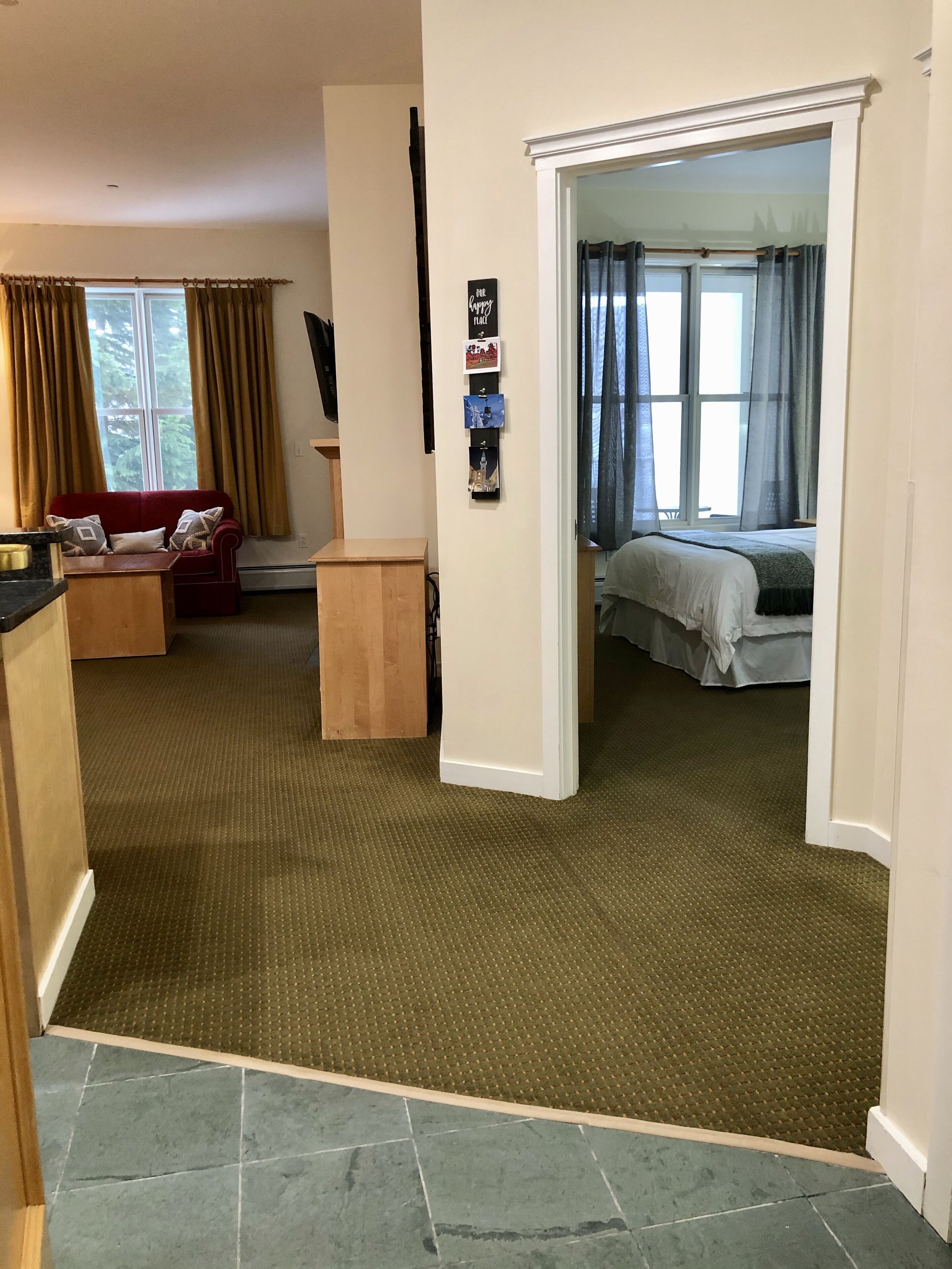 Un salon avec un canapé rouge, une table basse en bois et une grande fenêtre avec des rideaux. Un couloir mène à une chambre à coucher qui contient un lit et une fenêtre également dotée de rideaux.