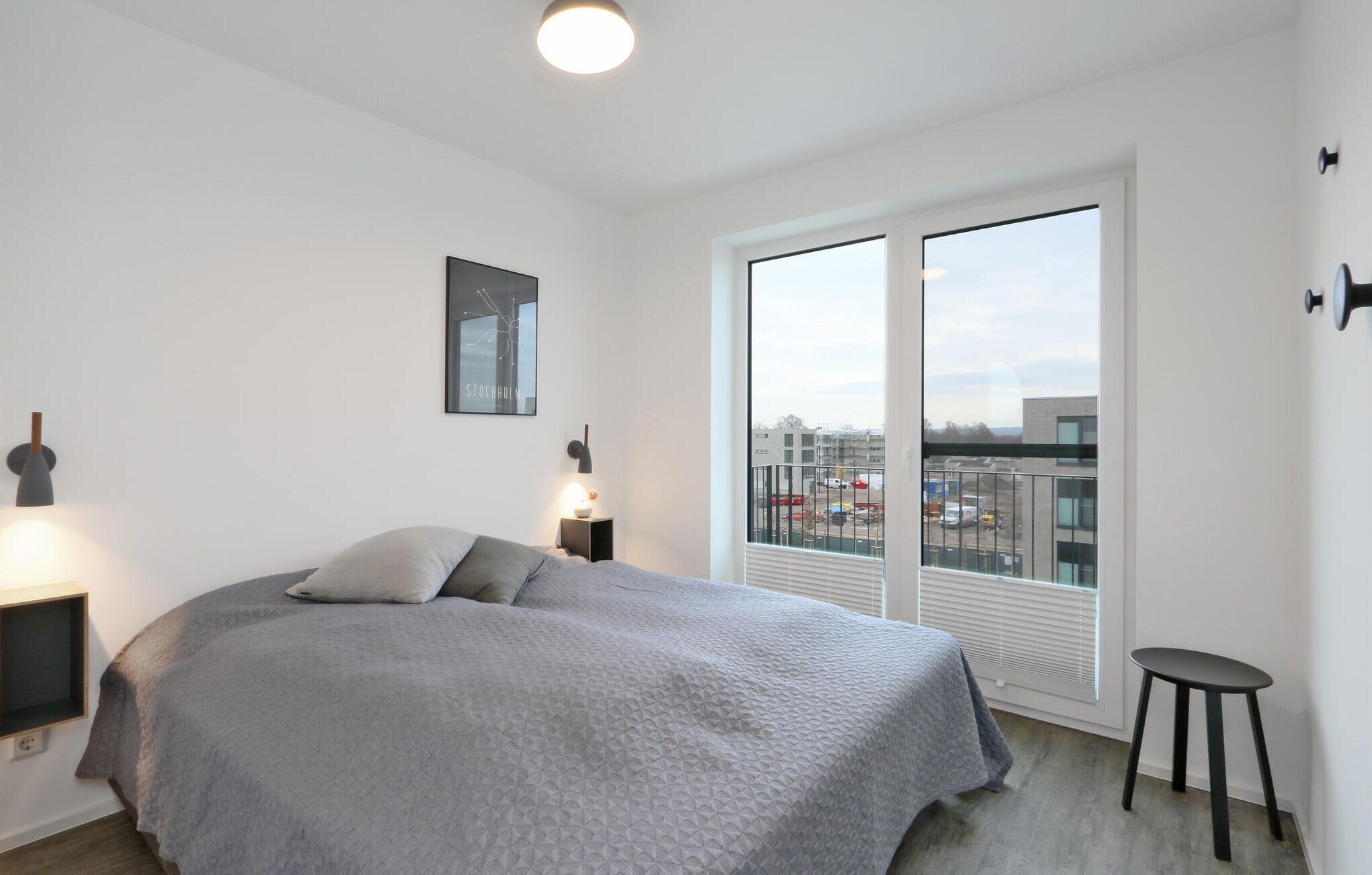 A modern bedroom with a large bed, a nightstand, a wall-mounted lamp, a framed picture, and a view of the city through large windows.