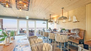 A spacious dining area with a view of the sea, featuring wooden ceiling beams, pendant lighting, and a bar area with stools. - Nice apartment in Lembruch/Dümmer See with indoor swimming pool (Lembruch/Dümmer See)