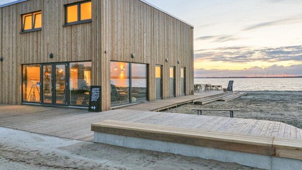 A modern wooden building with large windows, a wooden deck, and a view of the sea. - Nice apartment in Lembruch/Dümmer See with indoor swimming pool (Lembruch/Dümmer See)