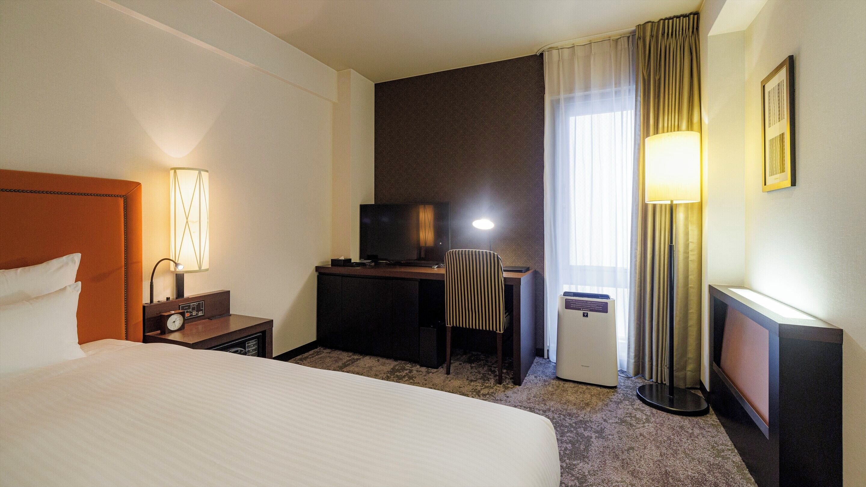 Une chambre d’hôtel avec un grand lit, un bureau avec une télévision, une chaise, une lampe de bureau, une horloge et un petit climatiseur.