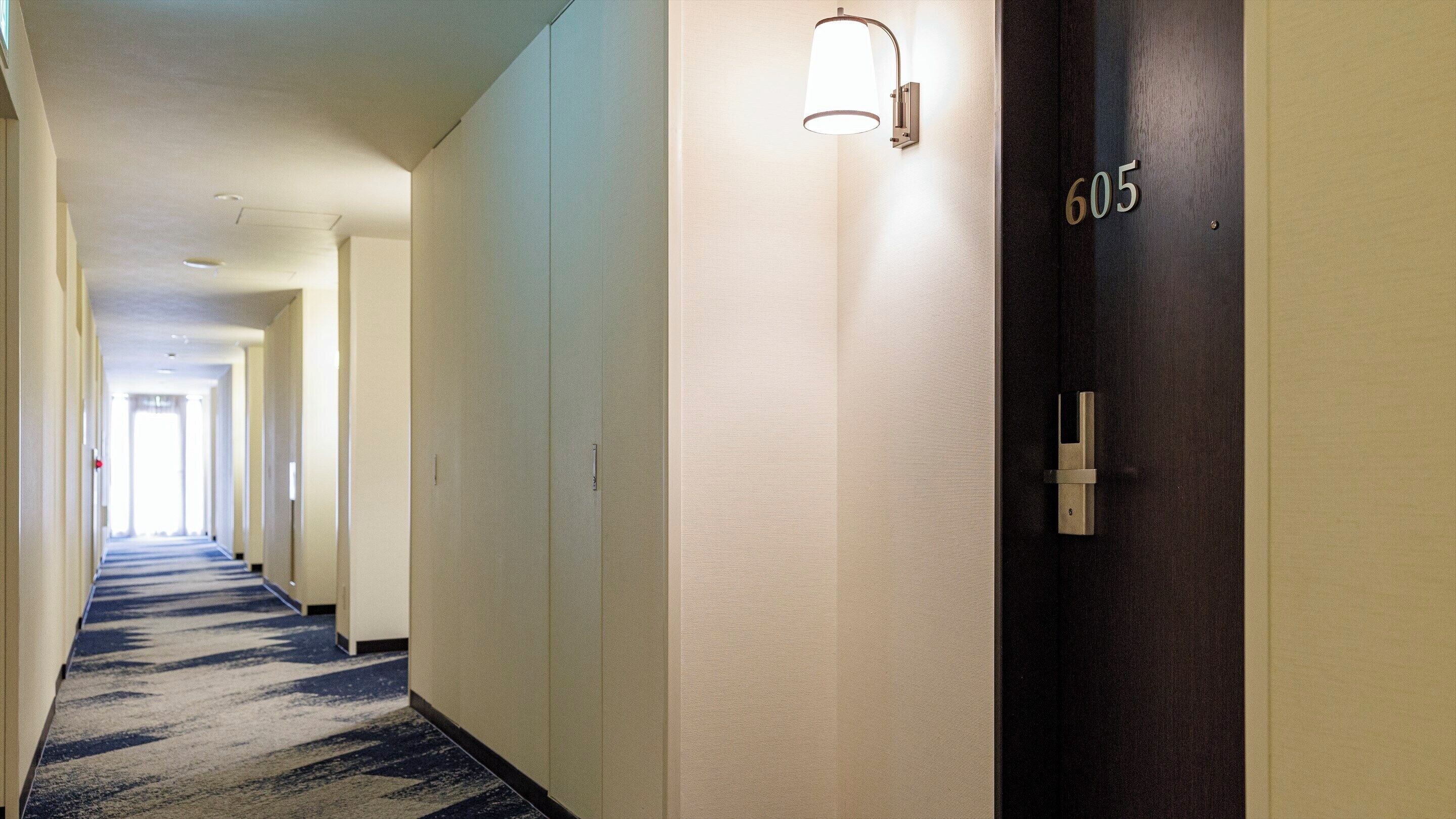 A hotel hallway with a carpeted floor, a wall-mounted light fixture, and a door with the number 605.