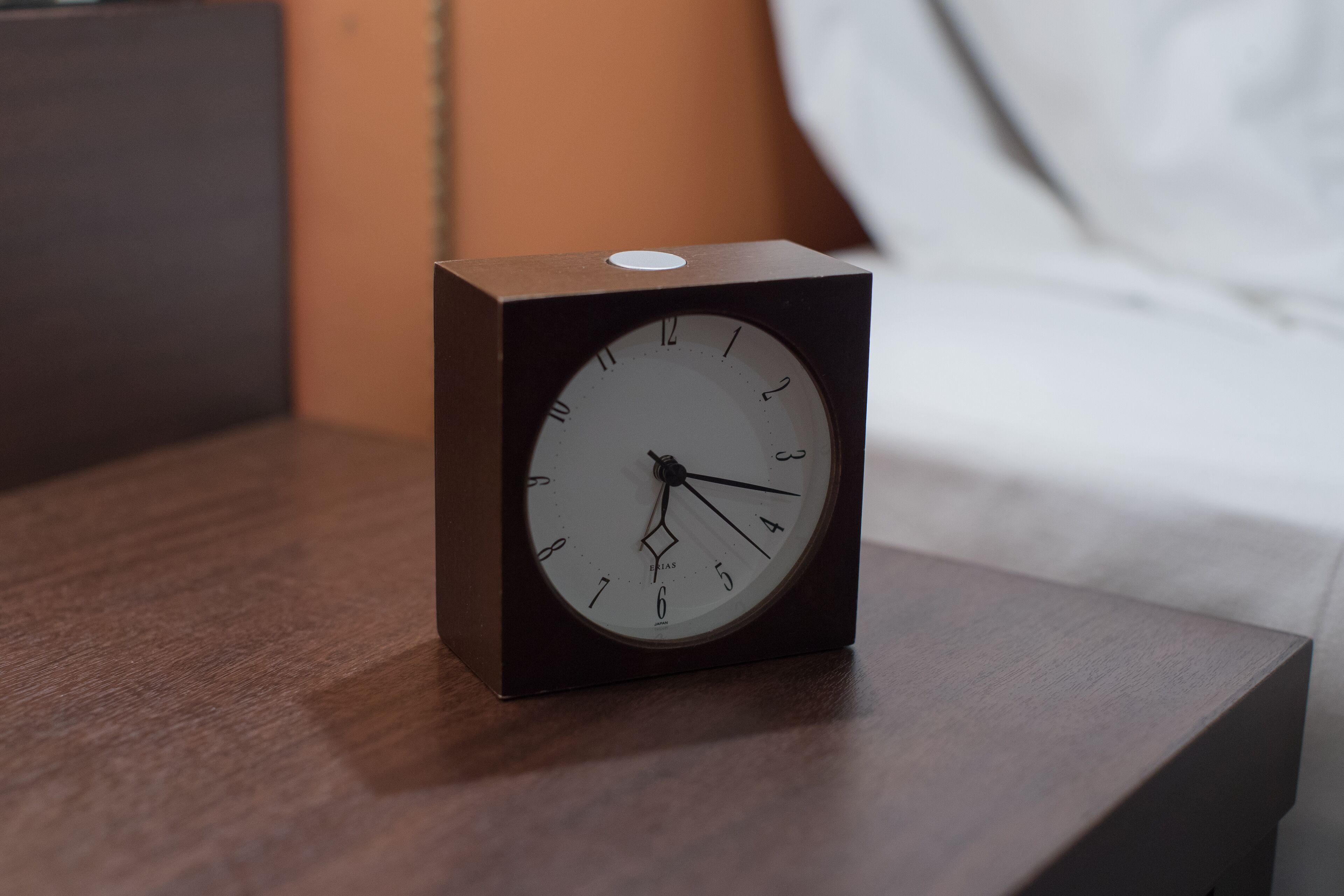 Una mesita de noche cuadrada, de madera oscura, con un reloj redondo y blanco que marca la hora.