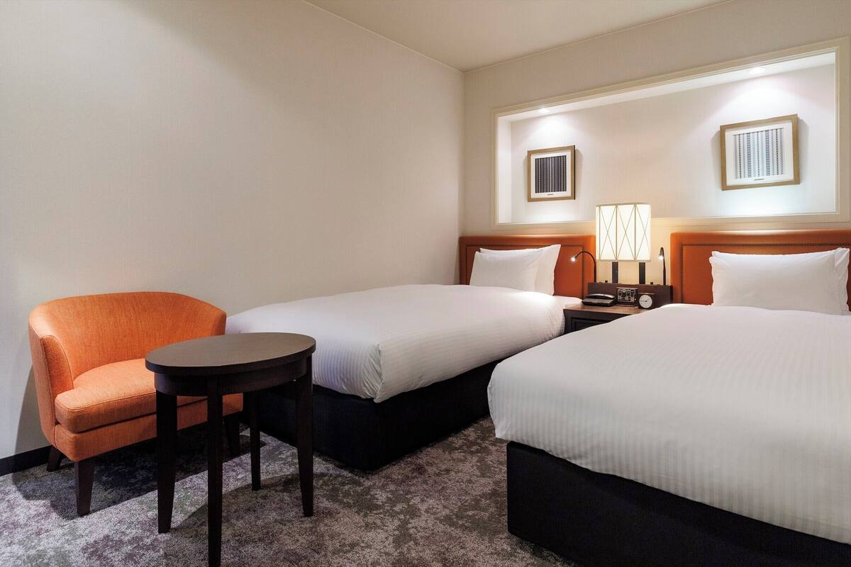 A hotel room with two single beds, a small table, a chair, a nightstand with a lamp, and framed pictures on the wall.