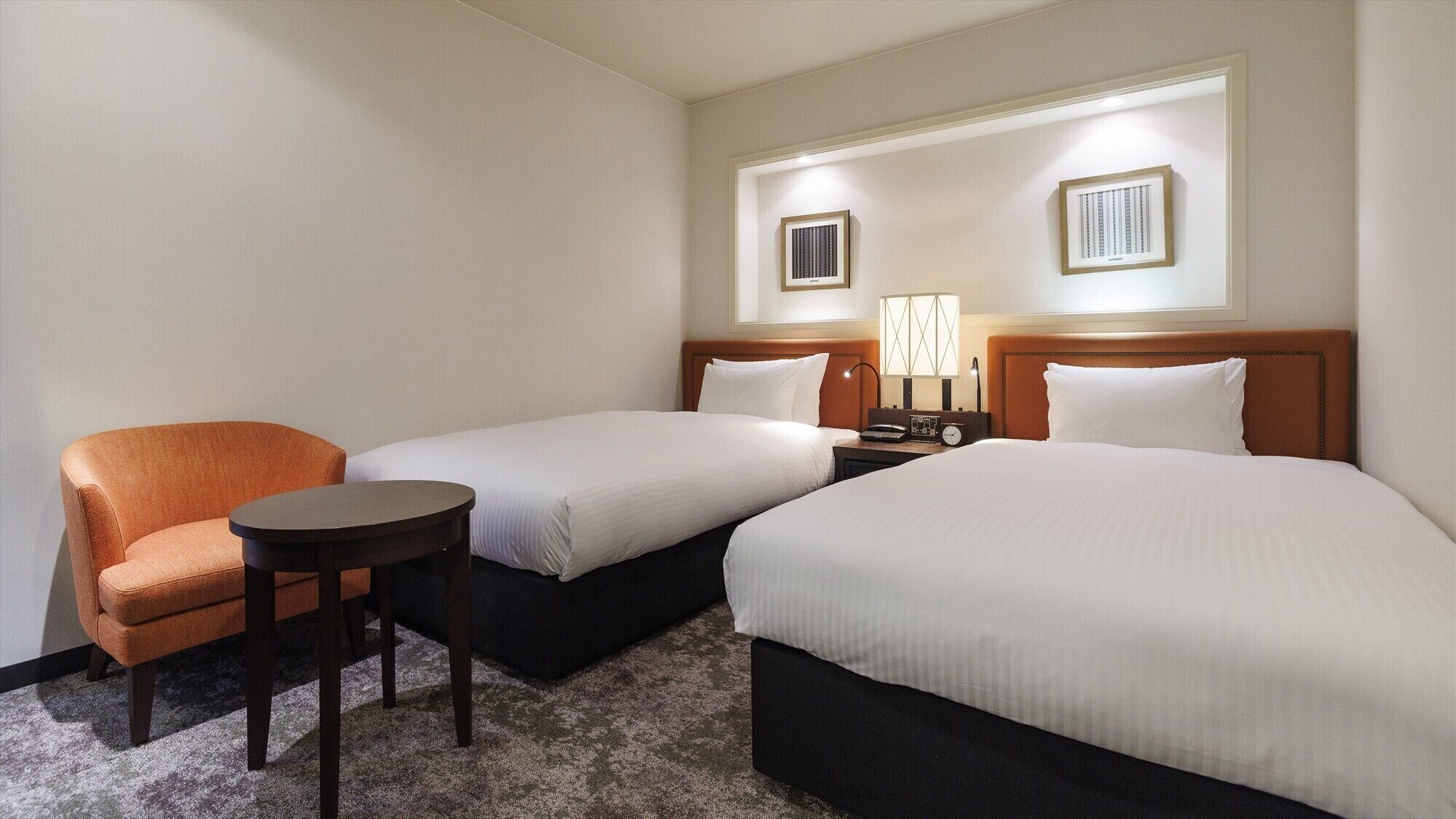 A hotel room with two beds, a chair, a small table, and a nightstand with a lamp.