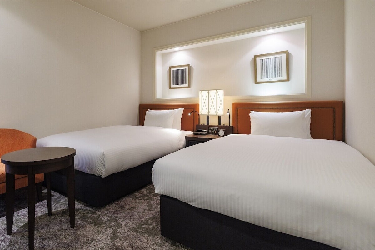 A hotel room with two beds, a chair, a small table, and a nightstand with a lamp.