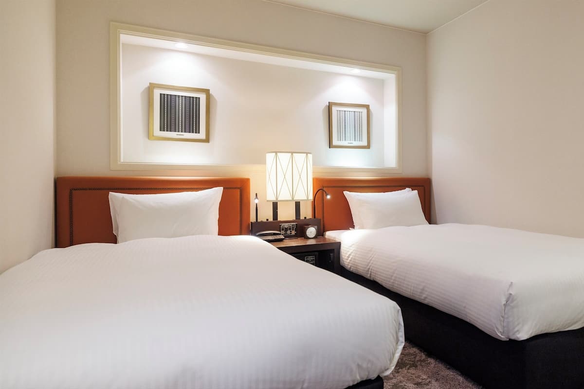 A hotel room with two single beds, a nightstand with a lamp, and two framed pictures on the wall.