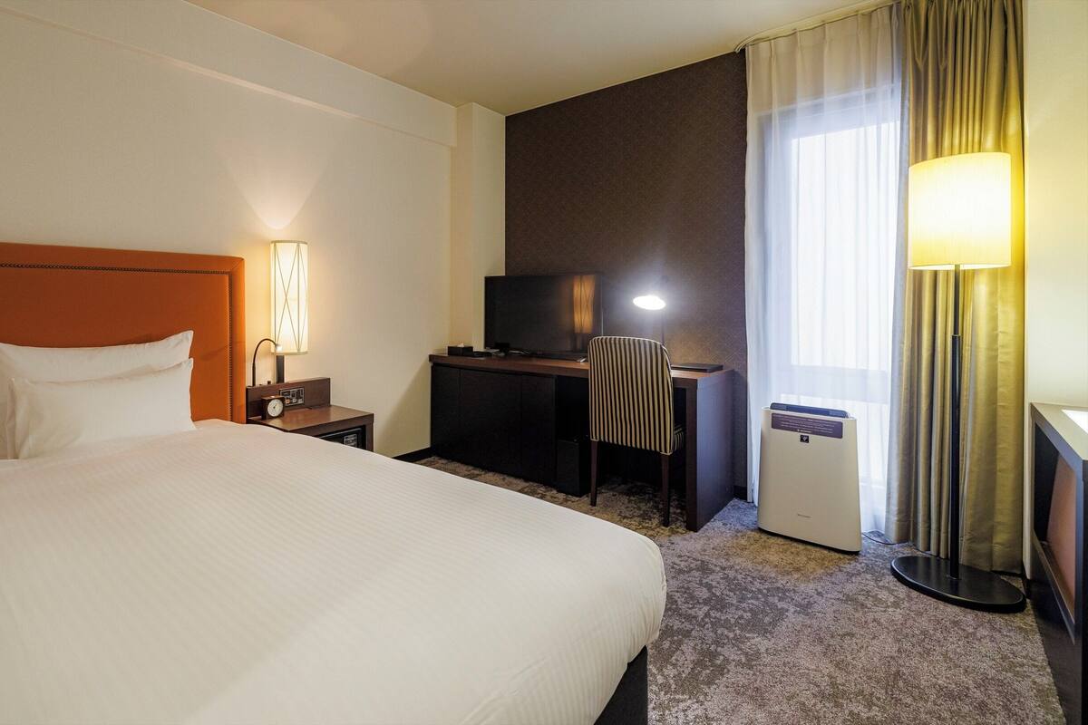 A hotel room with a large bed, a desk with a television, a chair, and a lamp.