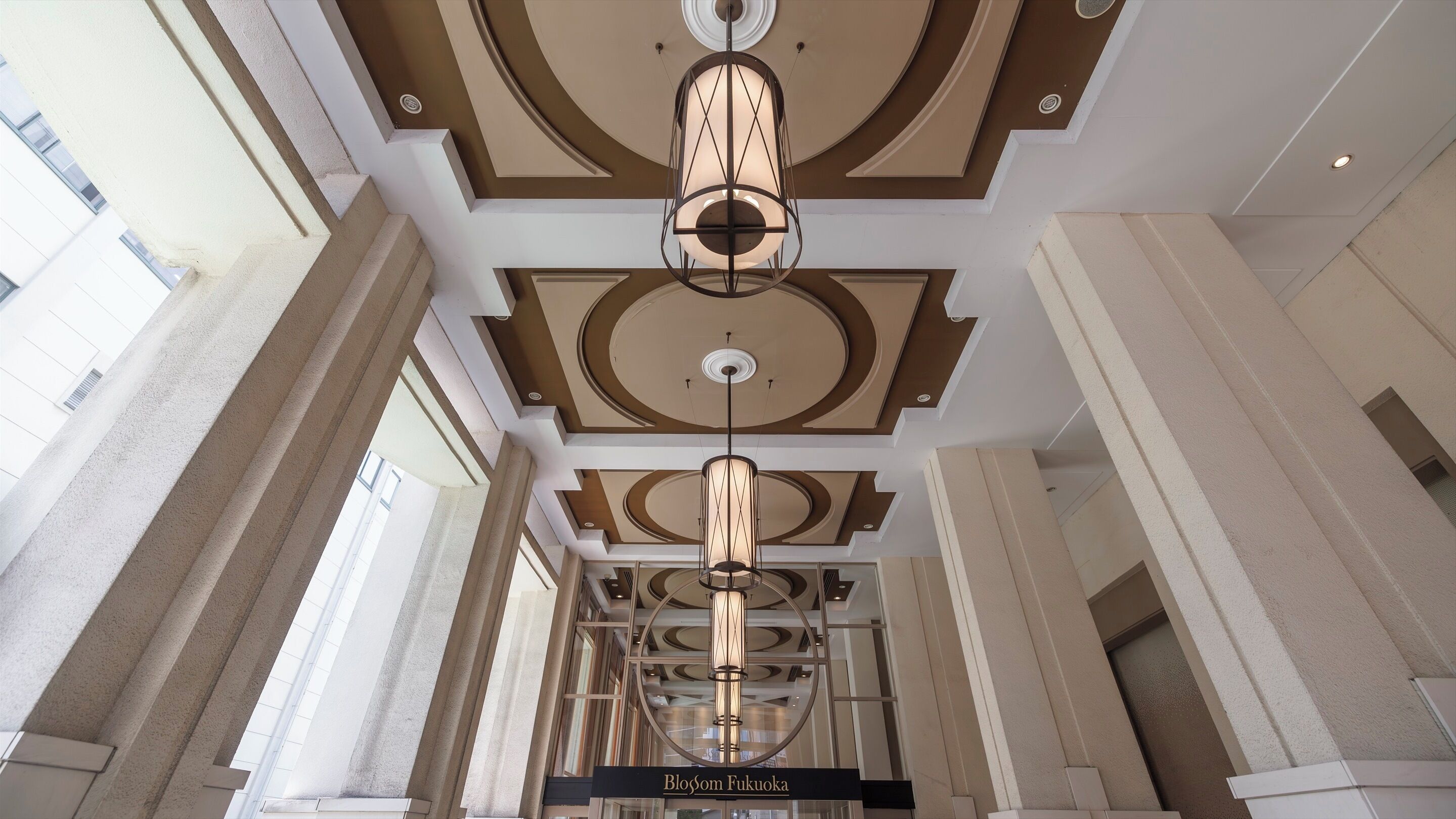 A hotel lobby with a central chandelier, high ceilings with decorative molding, and a sign reading "Blossom Fukuoka".