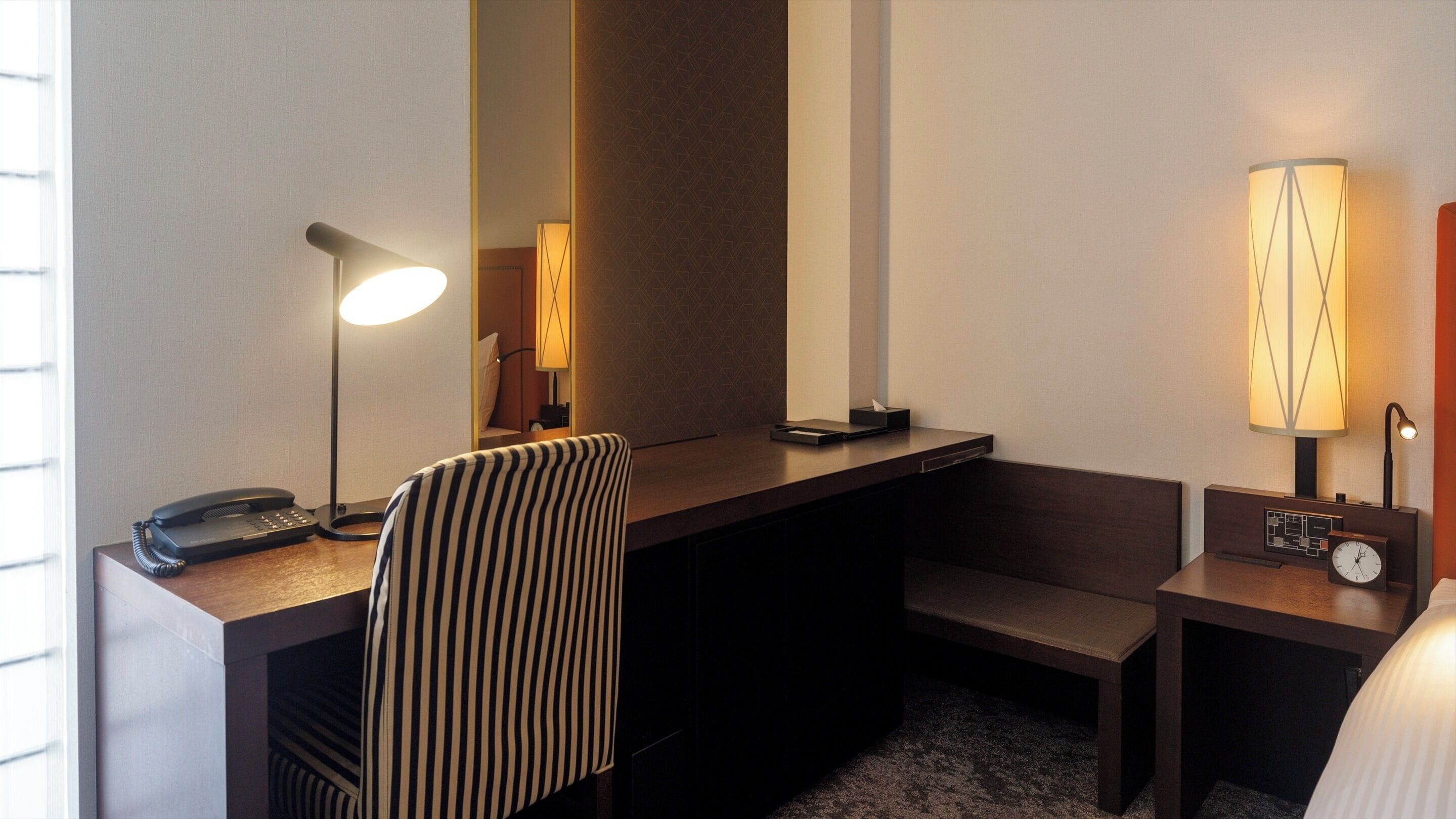 Une chambre d’hôtel équipée d’un bureau, d’un téléphone, d’une chaise à rayures, d’une table de chevet avec une lampe et d’une horloge.