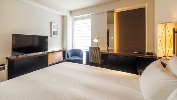 A hotel room with a large bed, a flat-screen TV, a desk with a chair, a lamp, and a large mirror.