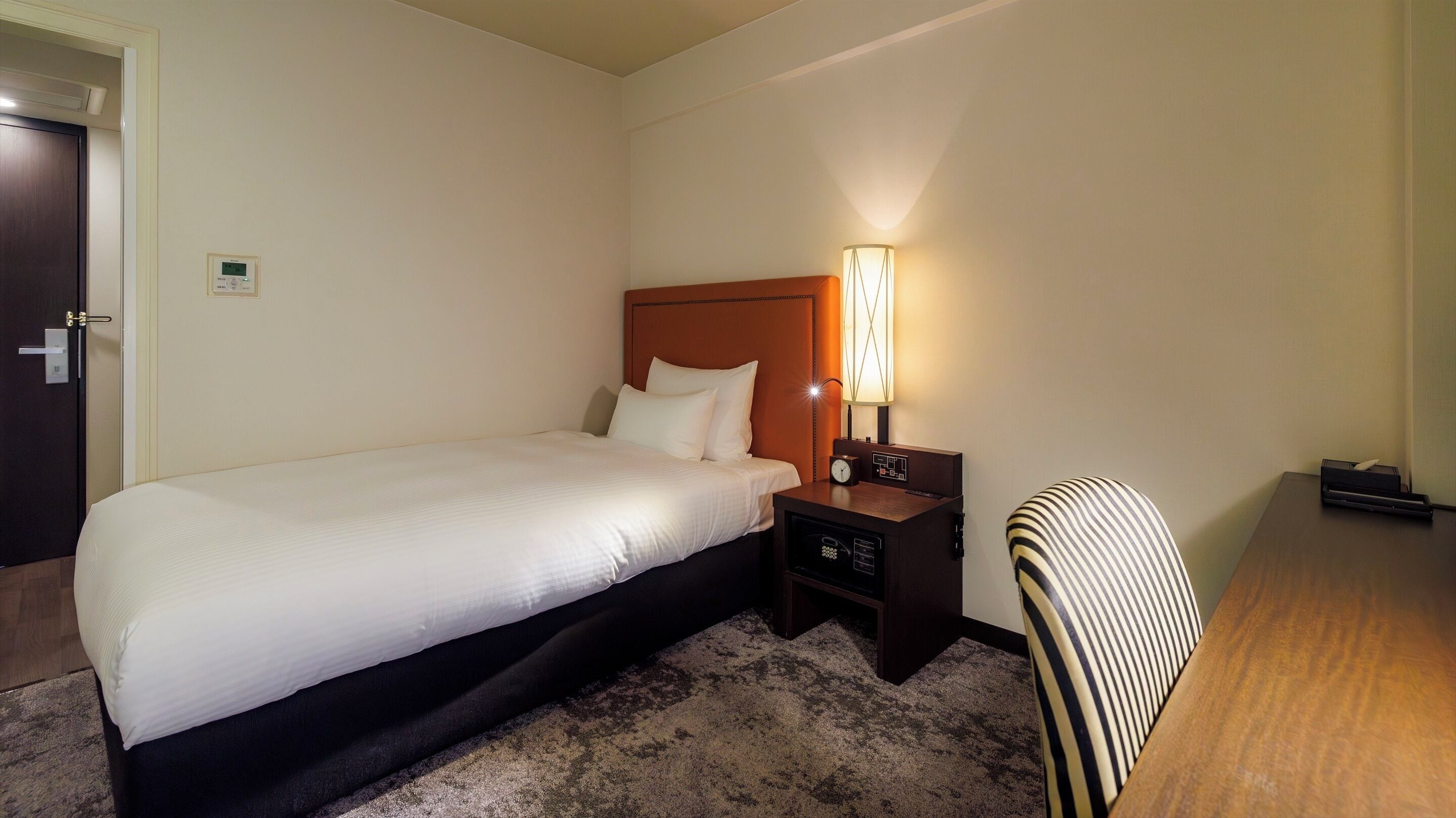 A hotel room with a single bed, a nightstand with a lamp, a chair, and a desk with a computer.