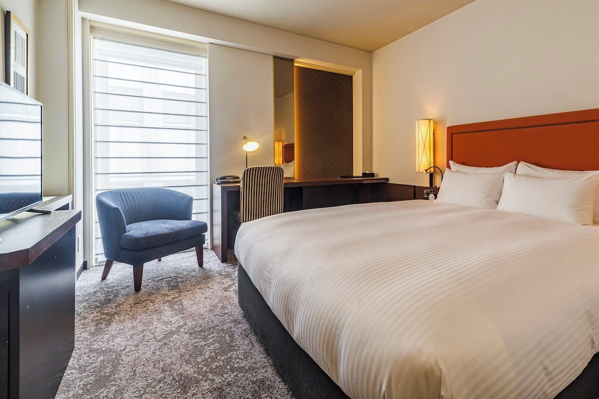 A hotel room with a large bed, a desk with a chair, a TV, and a window with blinds.