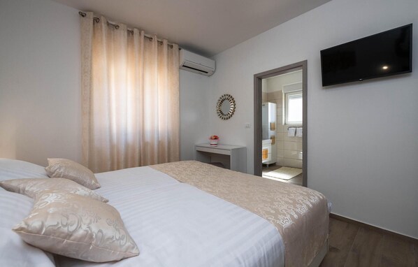 A hotel room with a bed, a TV mounted on the wall, an air conditioning unit, and a mirror reflecting a bathroom with a bathtub and towel rack.