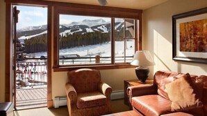 Ein gemütliches Wohnzimmer mit großem Fenster und Blick auf die Berge, eine Ledersofa, ein gemustertes Sessel und ein kleiner Tisch mit Lampe.