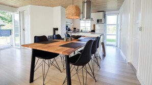 Uma ampla área de jantar com mesa de madeira e cadeiras pretas, luminária pendente e cozinha ao fundo.
