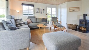 Uma sala de estar aconchegante com um sofá cinza, uma mesa de centro redonda e uma poltrona de vime. Há uma lareira e uma janela grande com vista para o exterior.