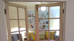 Ein Zimmer mit Holzboden, einem grünen Sessel, einem Bett mit Karomustern und einem farbenfrohen Sofa mit einem Kissen mit der Aufschrift 'Life is Beautiful'. Die Wand ist mit Fotos dekoriert.