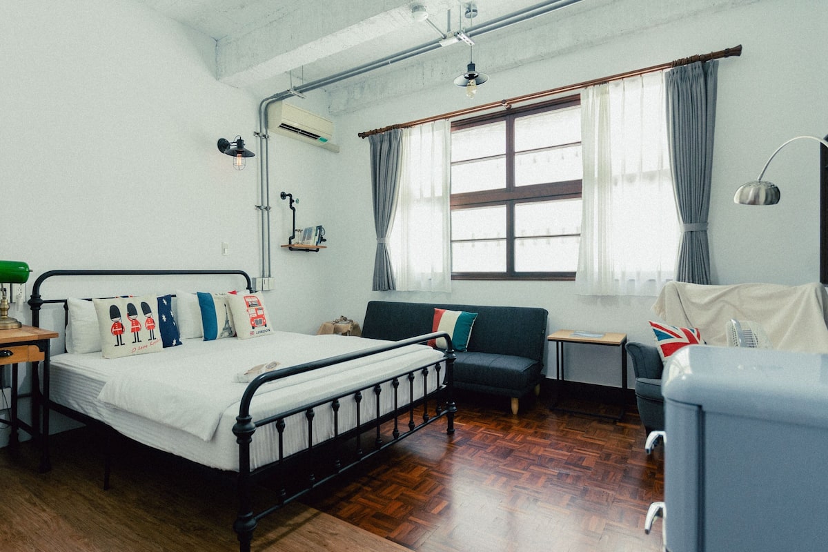 A bedroom with a bed, a sofa, a desk, and a chair. There is a window with curtains, a lamp, and a shelf with books.