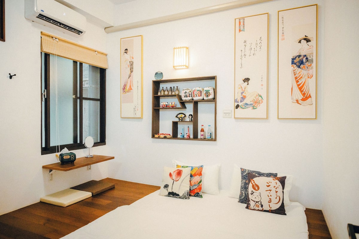 A neatly arranged bedroom with a wooden floor, a bed with white bedding, a shelf with decorative items, and traditional Japanese artwork on the walls.