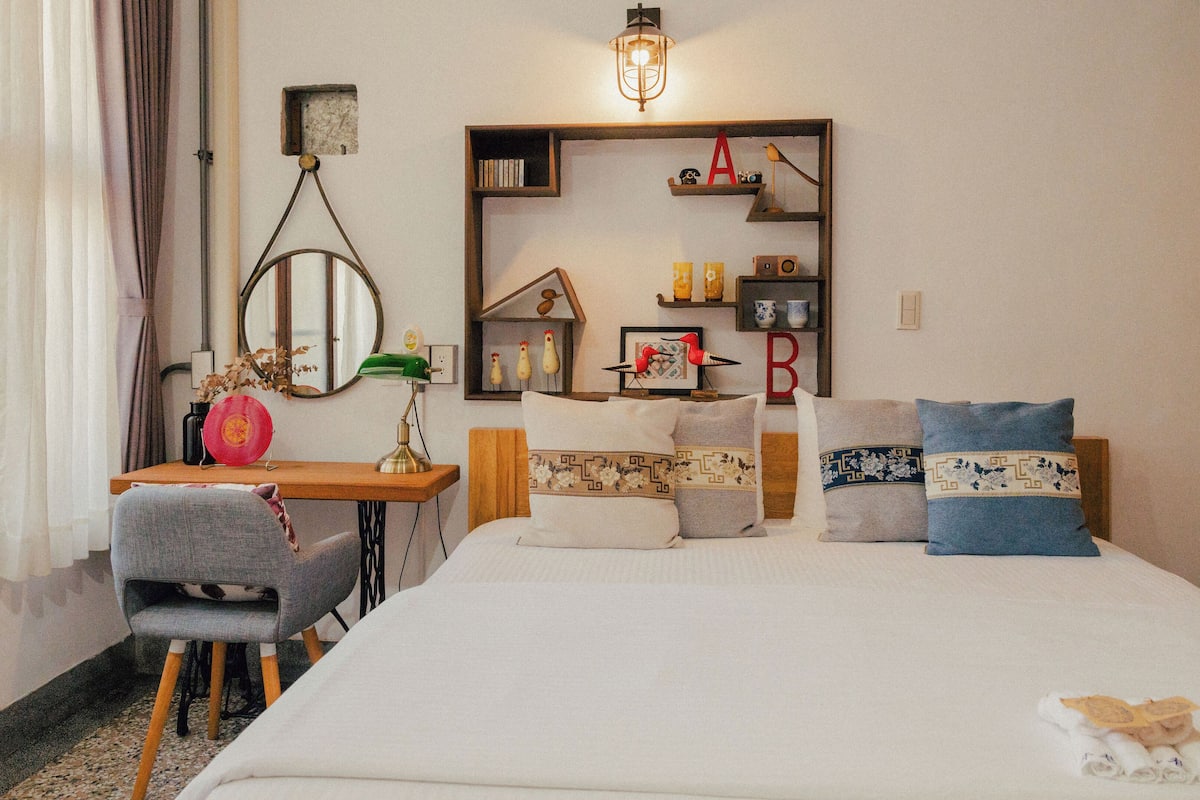A neatly arranged bedroom with a bed, a desk, a chair, and a shelf with decorative items.