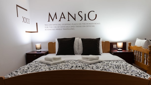 A neatly made bed with a patterned bedspread, two bedside tables with lamps, and a wall with a large Roman numeral and text. - Mansio (Niš)