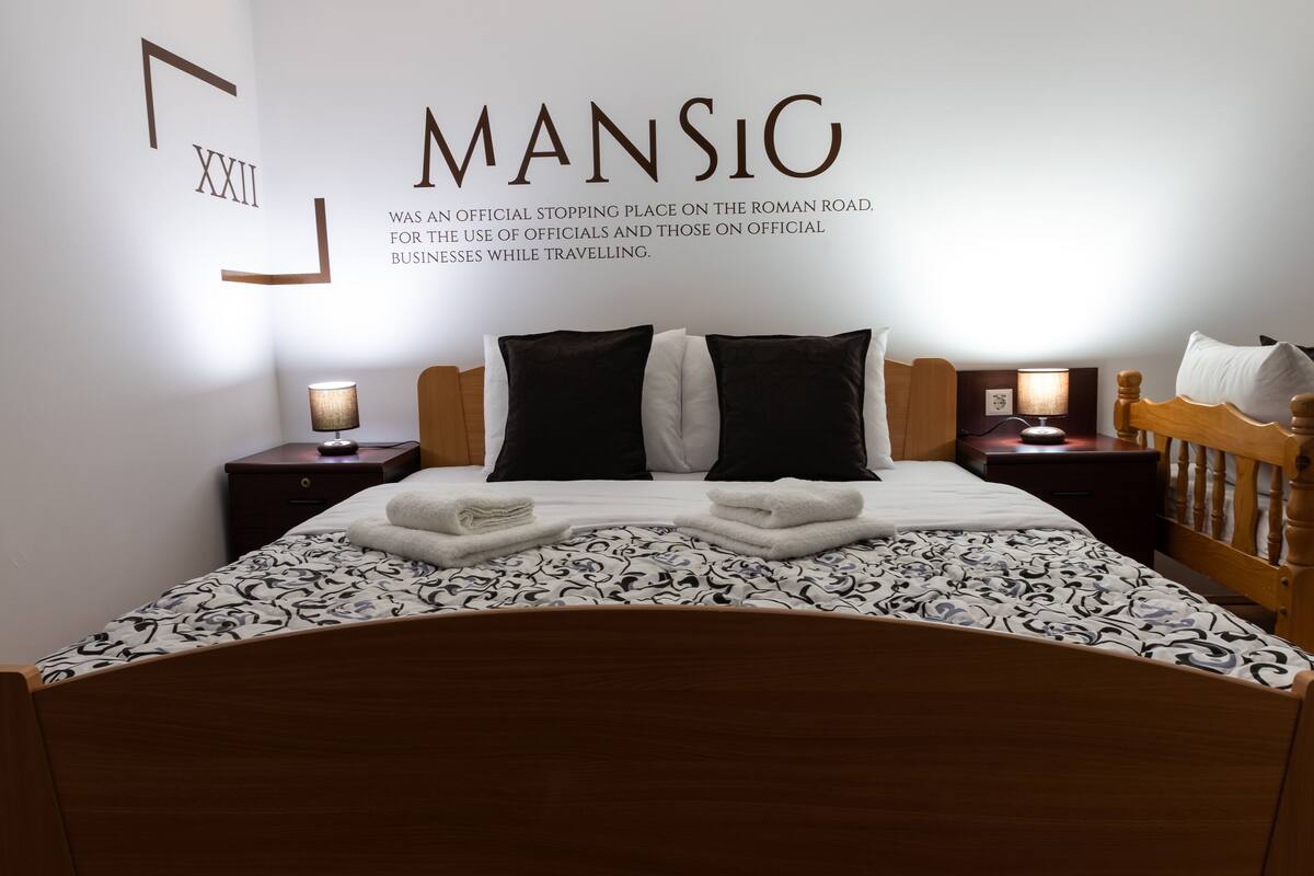 Una cama bien hecha con una colcha estampada, dos mesitas de noche con lámparas, y una pared con un gran número romano y texto.