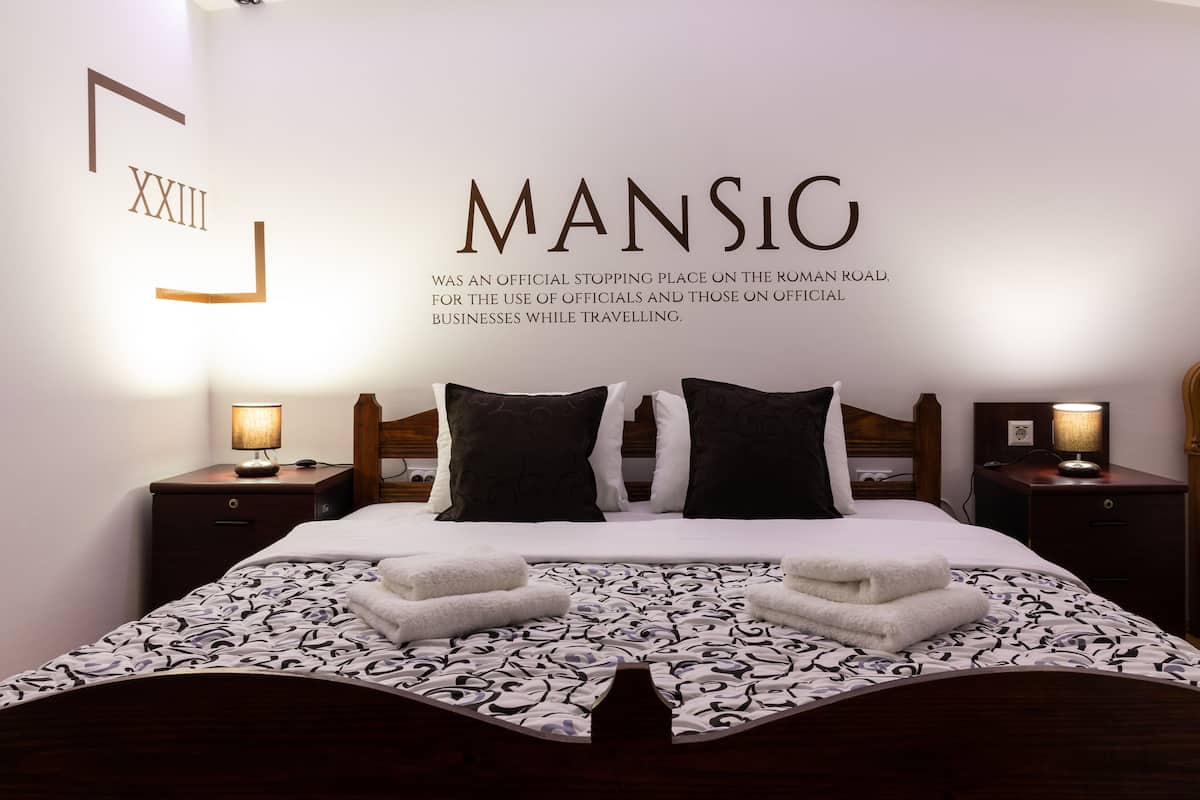 Una cama bien hecha con una colcha estampada, dos mesitas de noche con lámparas y una pared con un bloque de texto grande y otro pequeño.