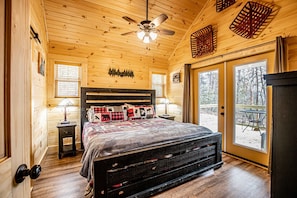 Une chambre confortable dans un chalet en rondins, dotée d’un grand lit, d’un ventilateur de plafond et offrant une vue sur l’extérieur grâce à une porte-fenêtre coulissante.