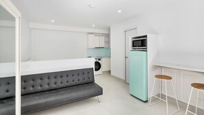 Un espace de vie moderne et compact, avec un canapé gris, un réfrigérateur de couleur turquoise et un petit coin cuisine équipé d’un lave-linge.