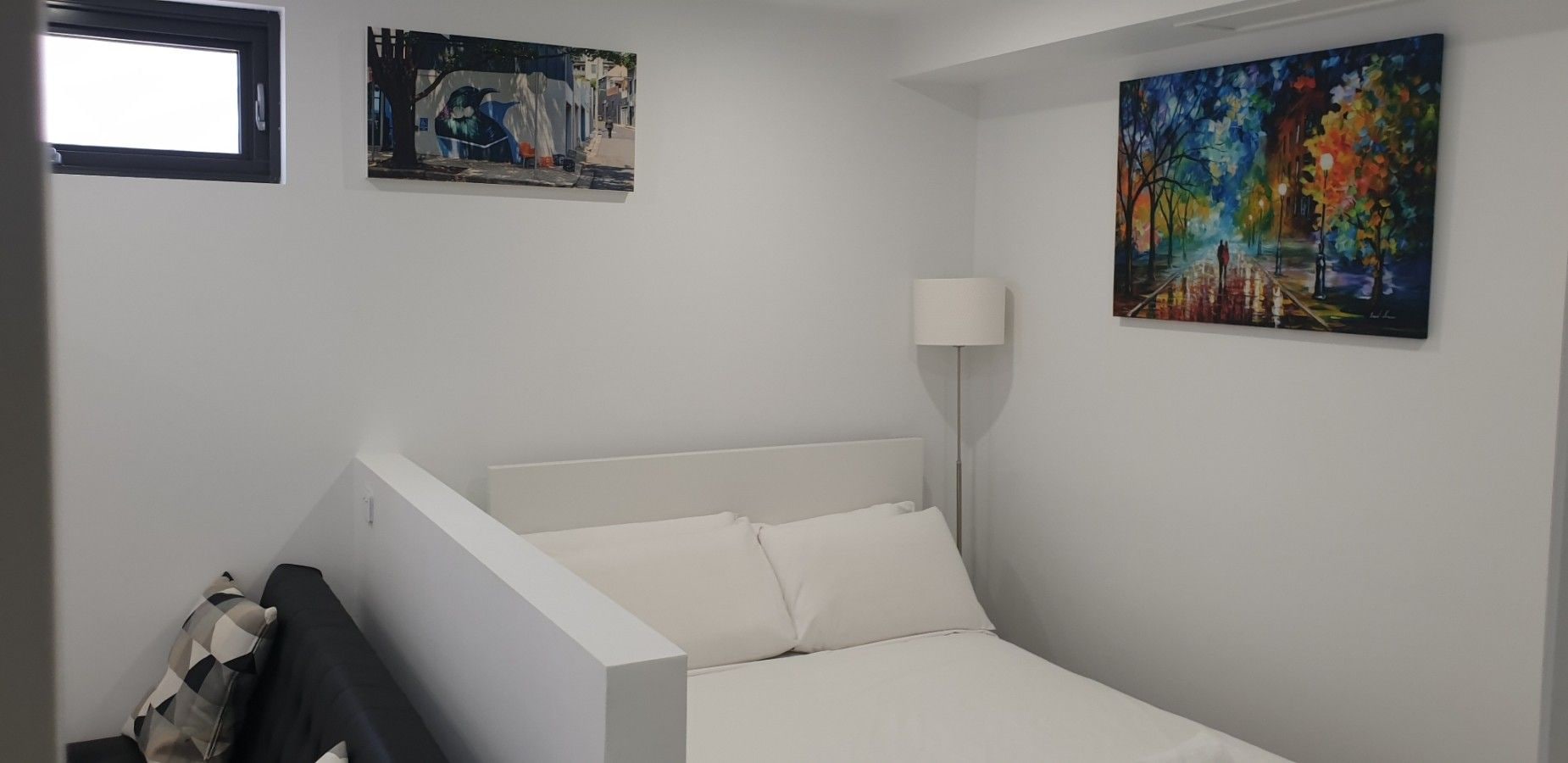 Een slaapkamer met een wit bed, een nachtkastje, een lamp en twee schilderijen aan de muur.
