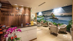 A hotel reception area with a wooden reception desk, a mural of a scenic landscape, and a seating area with chairs and a table. - Thanh Long Hotel Bach Dang (Ho Chi Minh City)