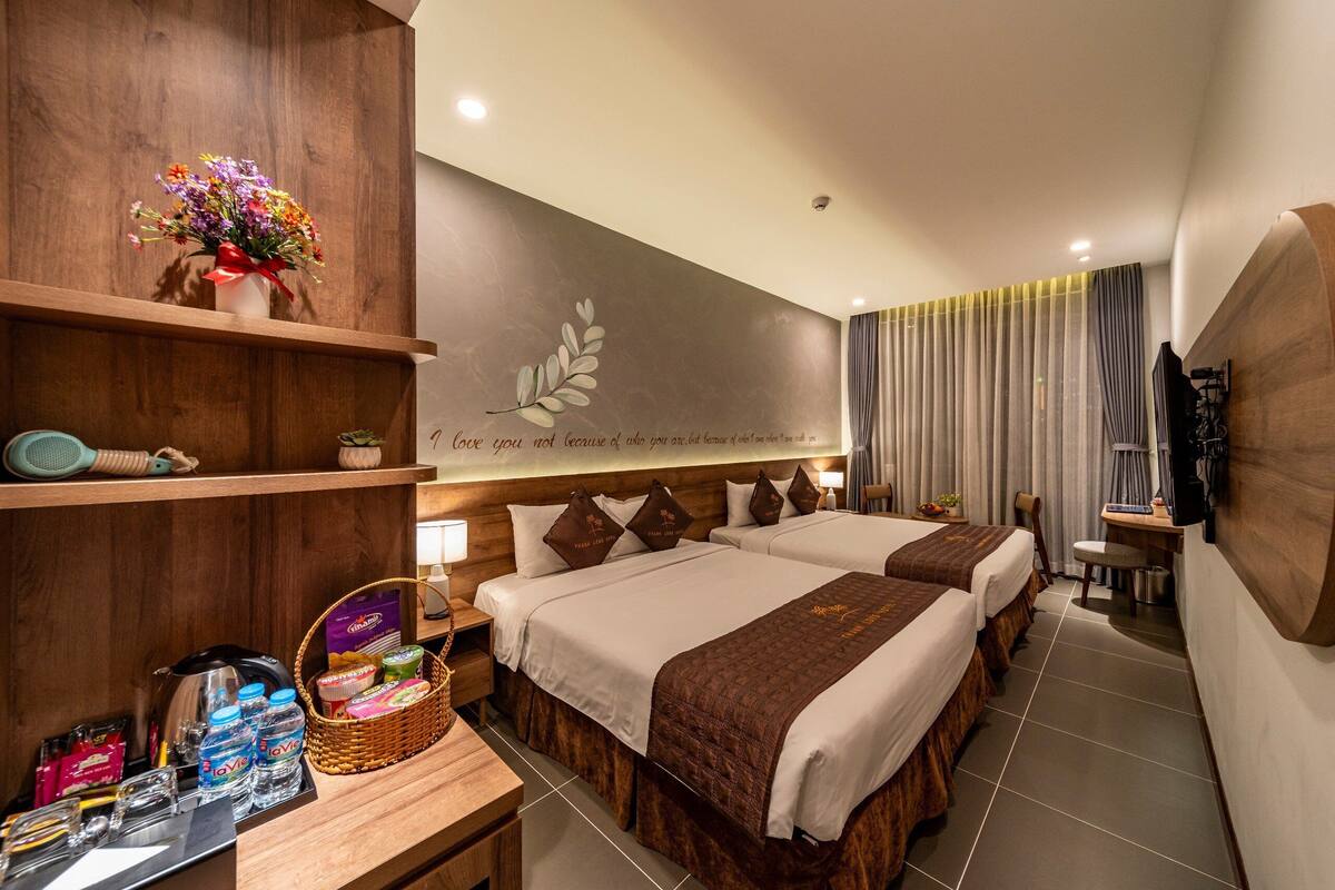 Ein Hotelzimmer mit zwei Betten, einer Holzablage mit einer Blumenvase, einem am Wand montierten Fernseher und einem Schreibtisch mit einem Tablett Toilettenartikel.