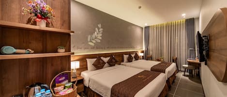 A hotel room with two beds, a wooden shelf with a vase of flowers, a wall-mounted TV, and a desk with a basket of toiletries.
