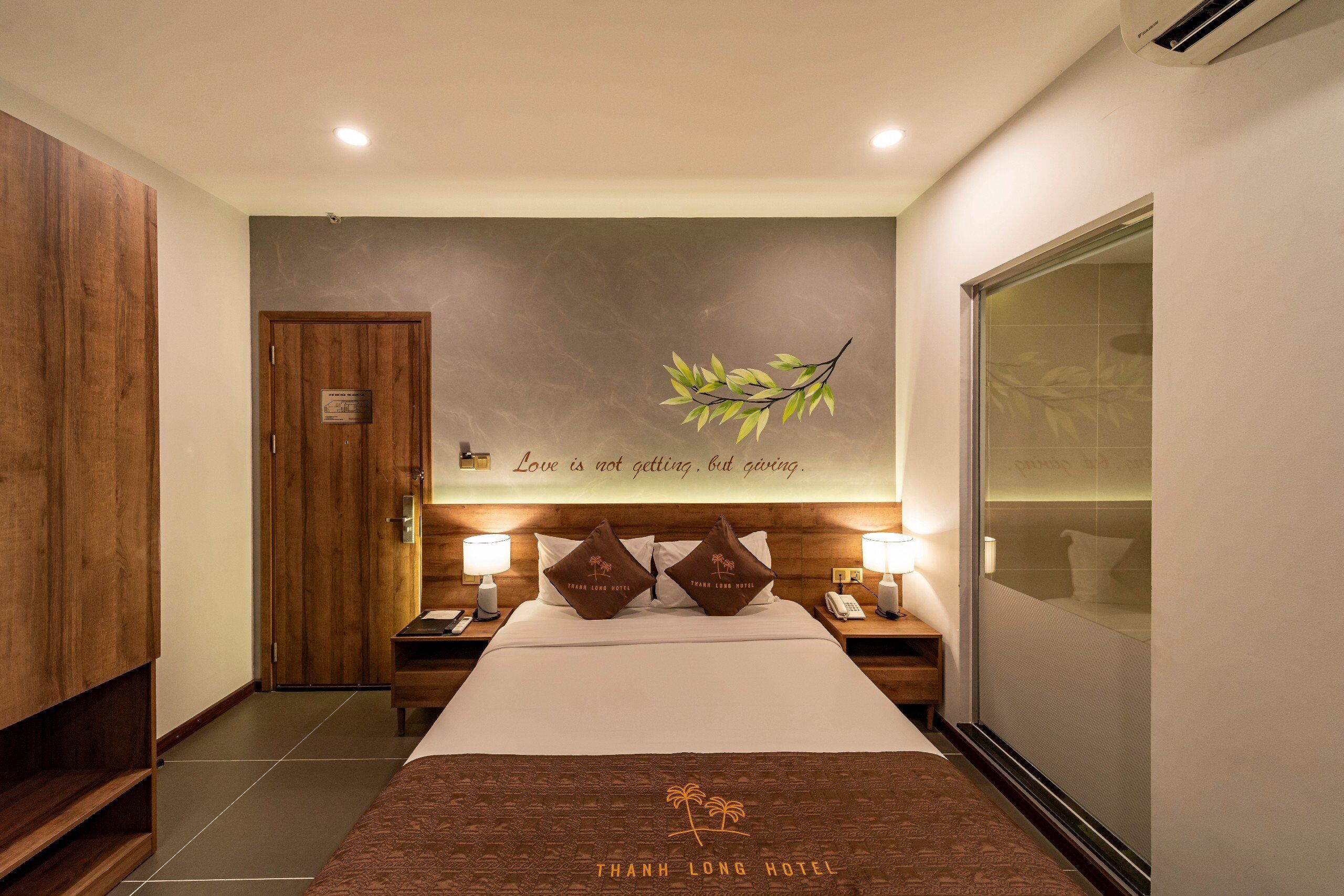 A hotel room with a bed, nightstands, a wooden wardrobe, a wall-mounted air conditioner, and a decorative leaf on the wall.