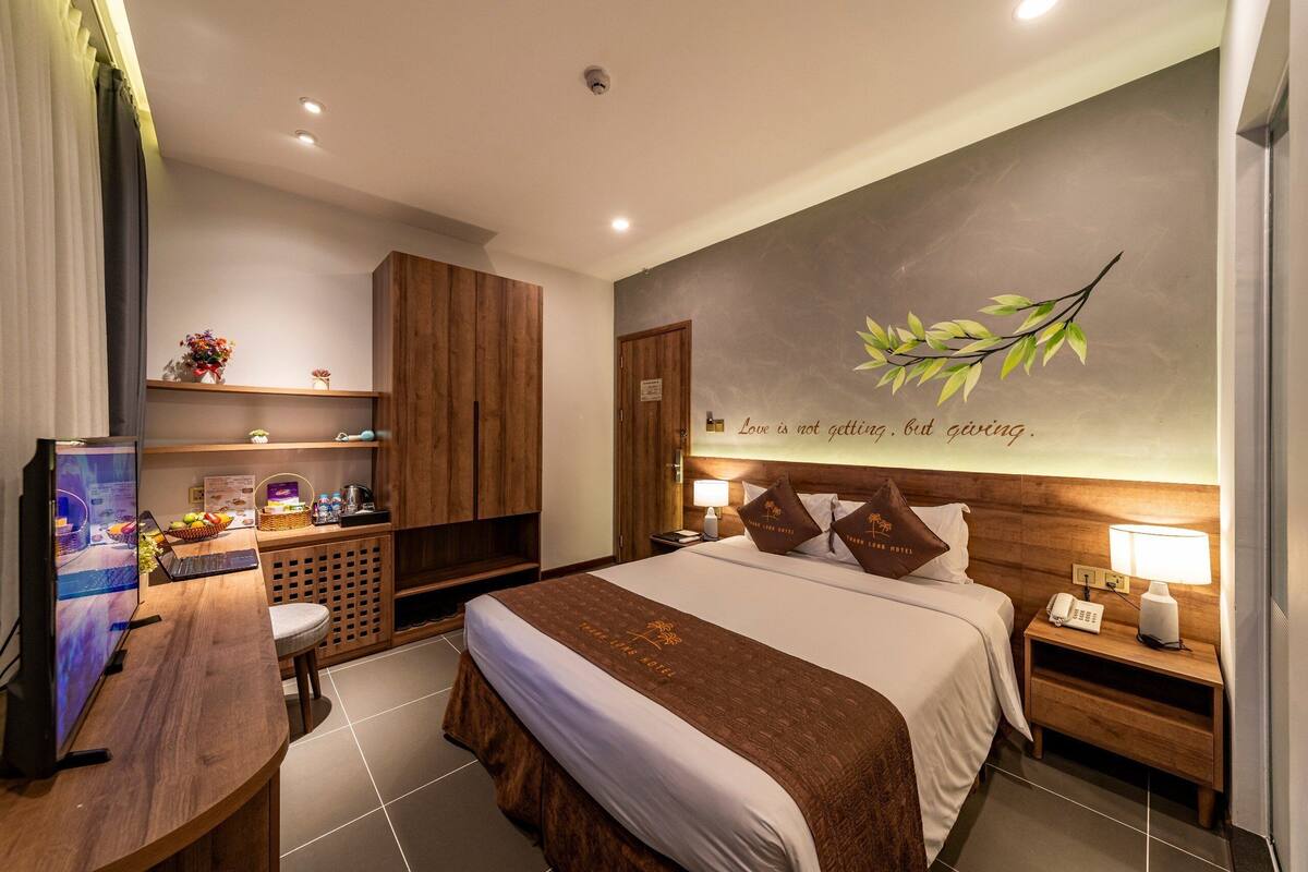 Ein Hotelzimmer mit einem großen Bett, einem Schreibtisch mit Computer, einer kleinen Sitzecke sowie einer Wand mit dekorativem Blattwerk und einem Motivationsspruch.