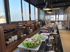 Un buffet proposant une variété de plats, notamment des salades et des plats chauds, dans un espace de restauration moderne doté de grandes fenêtres et d’un haut plafond.