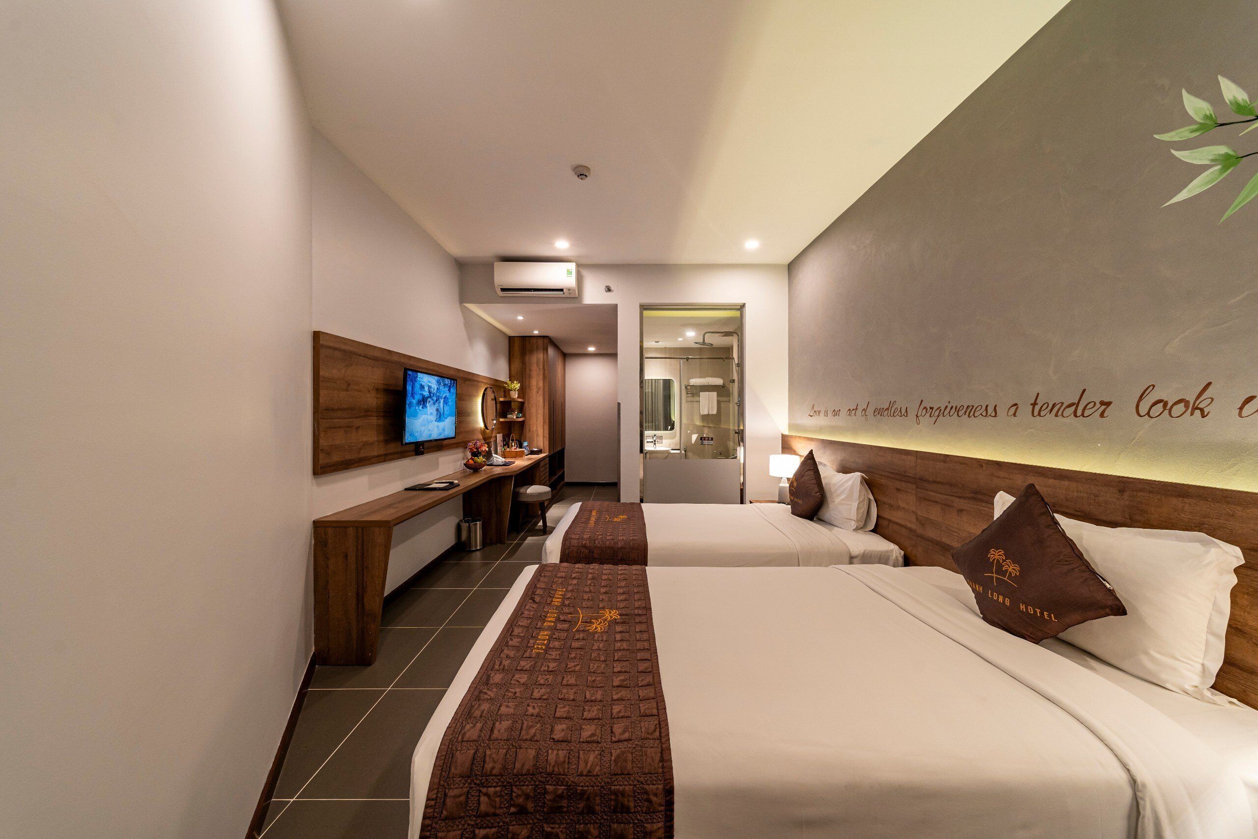 Habitación de hotel con dos camas, un escritorio con televisor, un baño y una pared decorada con una planta y texto.