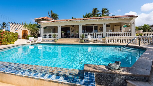 Vista de la piscina de un resort, con una piscina azul cristalina, una zona de estar cubierta y un edificio blanco con tejado de tejas.